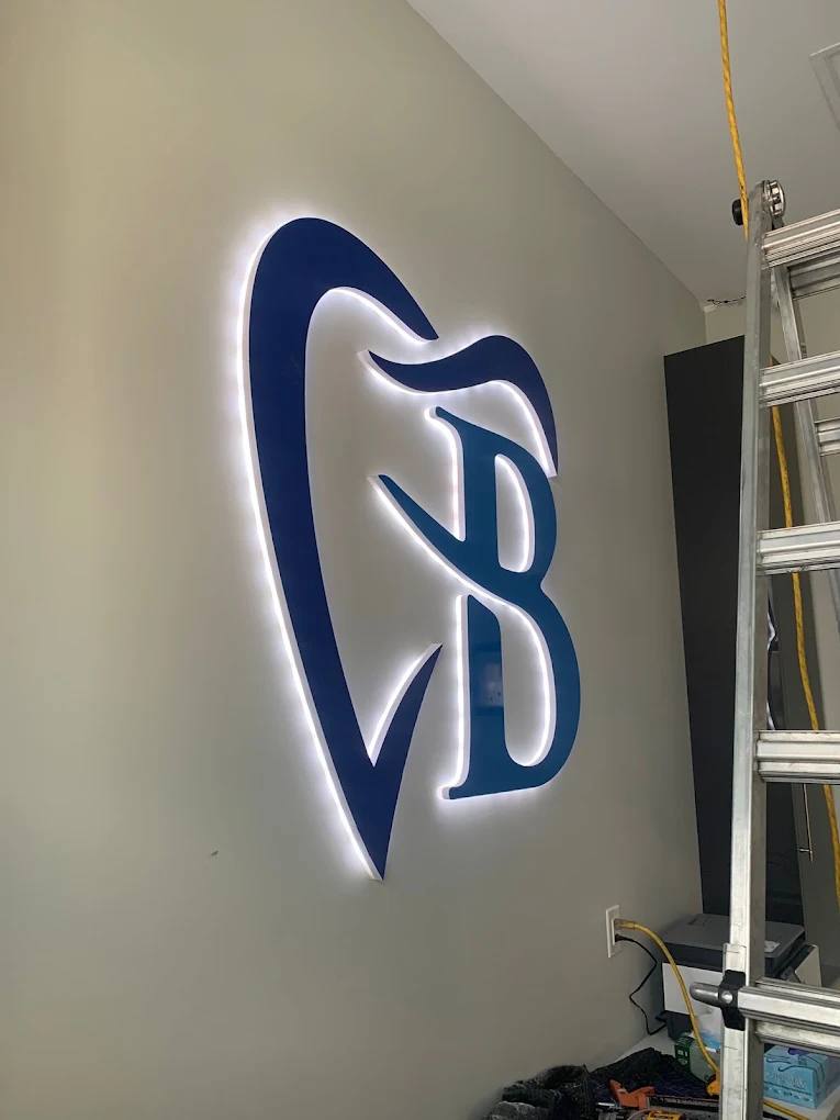 Blue dental clinic logo with a white halo-lit glow on a reception wall, ladder beside it during install